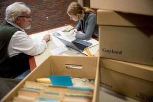 Phil Adamo studies at a table with a student. They are surrounded by boxes of files and papers.