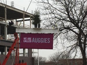 The beam, signed by members of the Augsburg community at the April groundbreaking ceremony, is hoisted into the air with a banner proclaiming, "We Are Called. Auggies." during the November 22, 2016, Topping Off ceremony. 