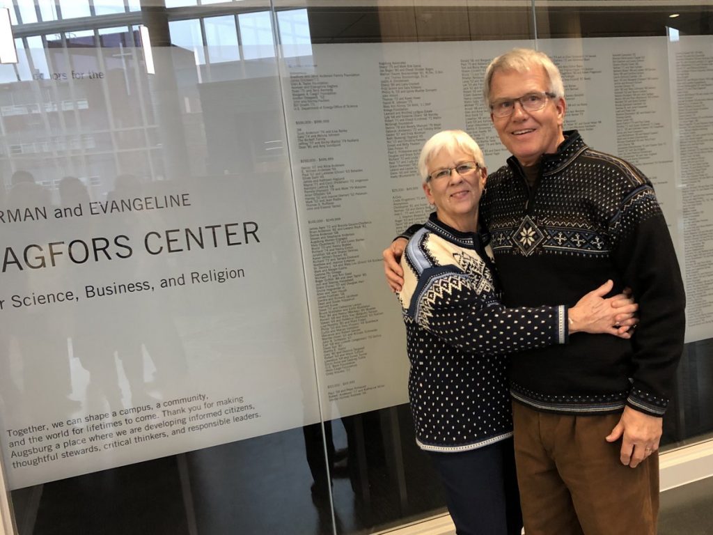 Man and woman wearing Norwegian sweaters in front of wall of donor names