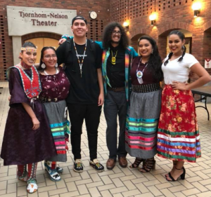 American Indian students wearing traditional attire stand outside the Tjornhom-Nelson Theater smiling next to one another