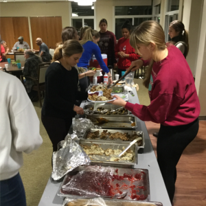 Students dish out food while others sit around a table.